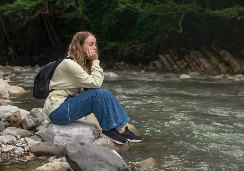 frustrated, tired tourist sits alone on the bank of a mountain river, depression, disappointment, despondency among young girls